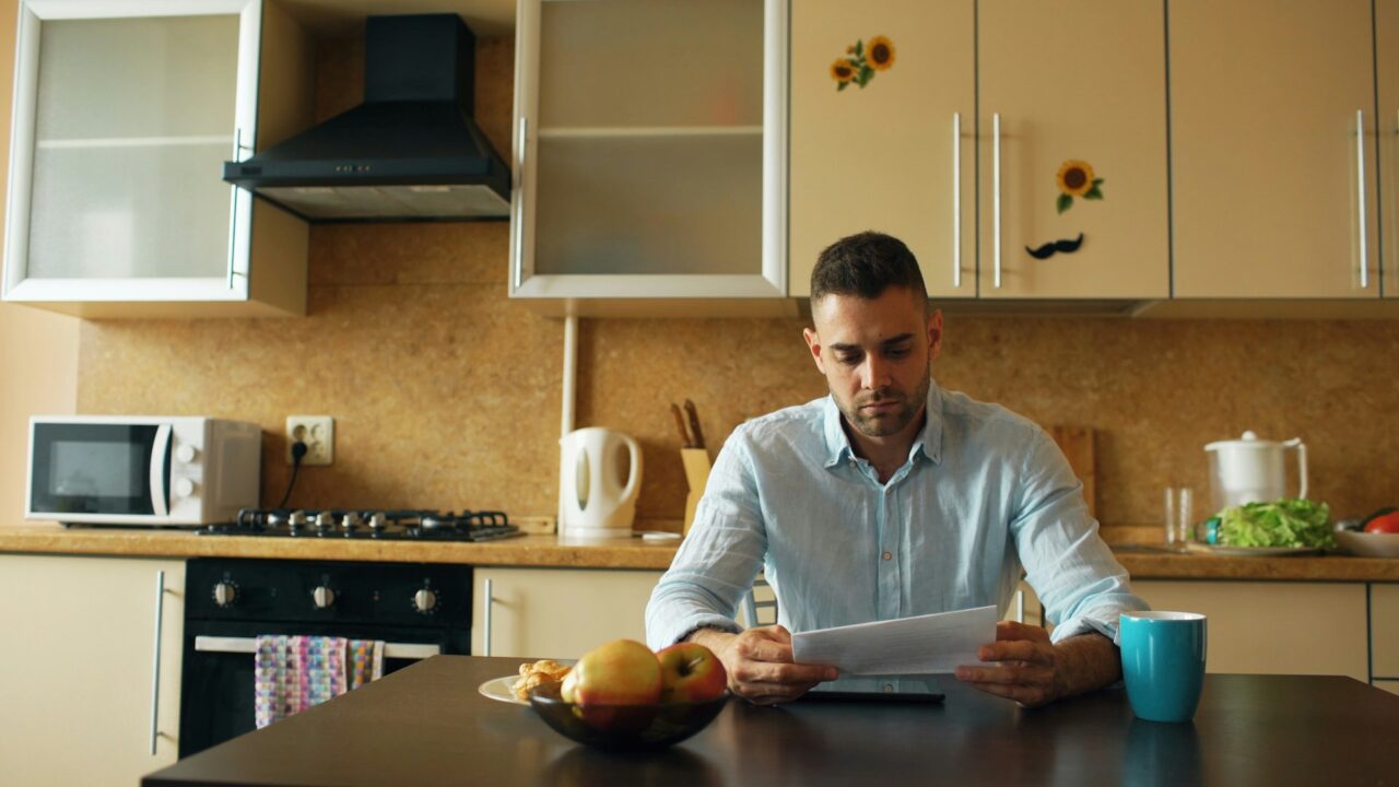 Man sitting at a kitchen table reading a document with a coffee cup nearby