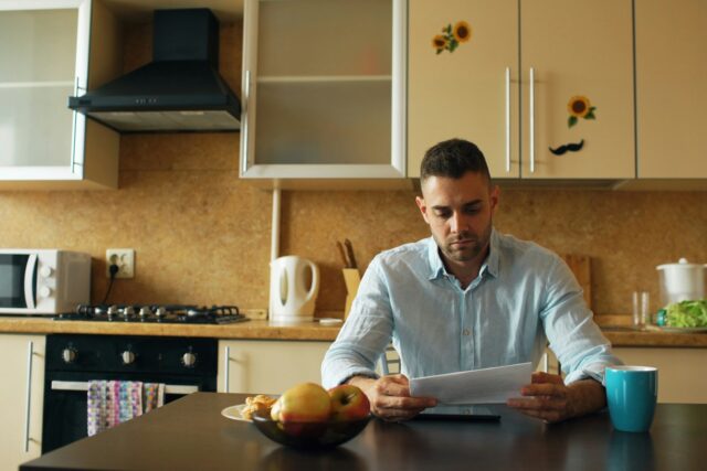 Man sitting at a kitchen table reading a document with a coffee cup nearby
