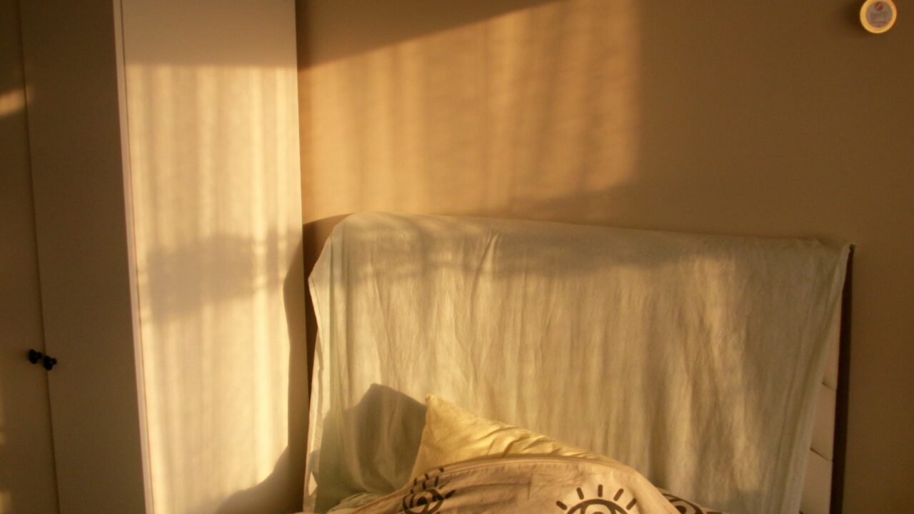 A simple bedroom with warm sunlight streaming through a window onto a bed with patterned bedding