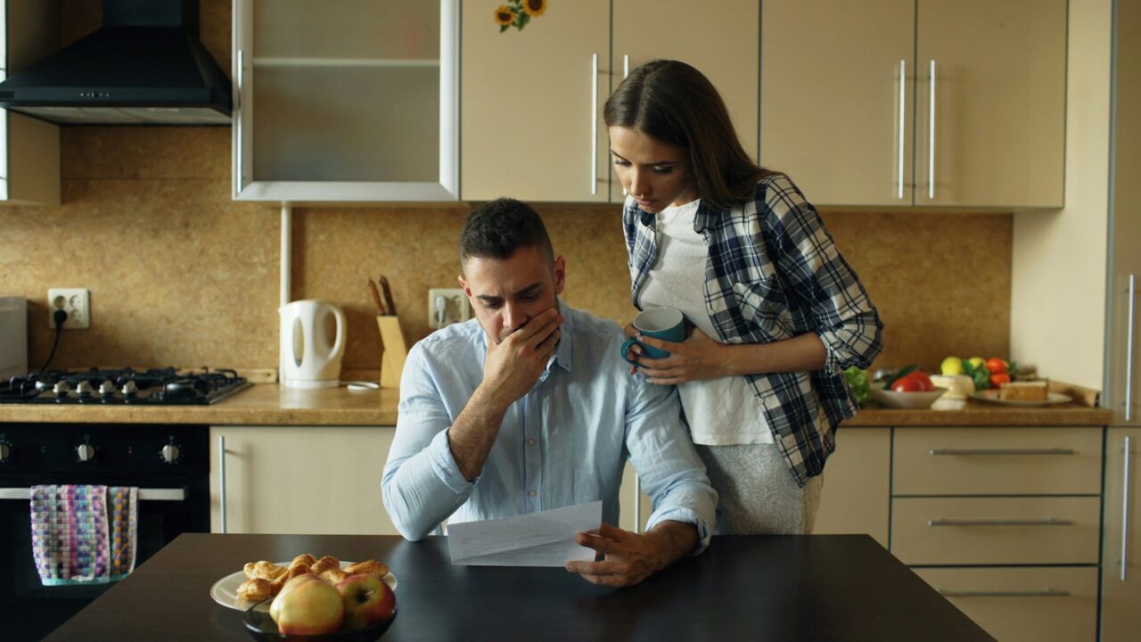 A man and woman at a kitchen table looking concerned while reviewing documents, with fruit visible on the surface.