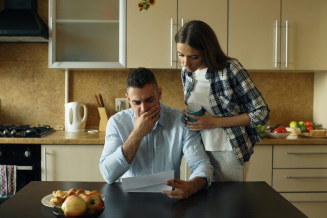 A man and woman at a kitchen table looking concerned while reviewing documents, with fruit visible on the surface.