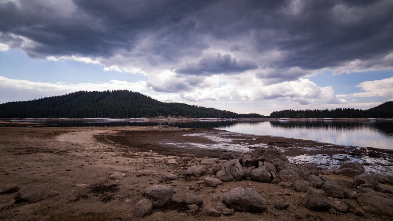 A large body of water with exposed muddy banks and dark cloudy sky above forested hills