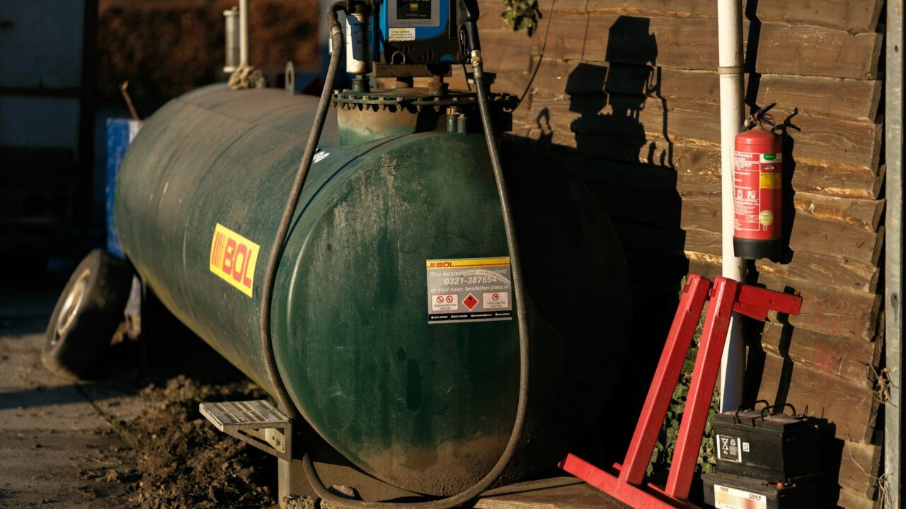A large green cylindrical heating oil tank positioned against a wooden building with safety equipment nearby