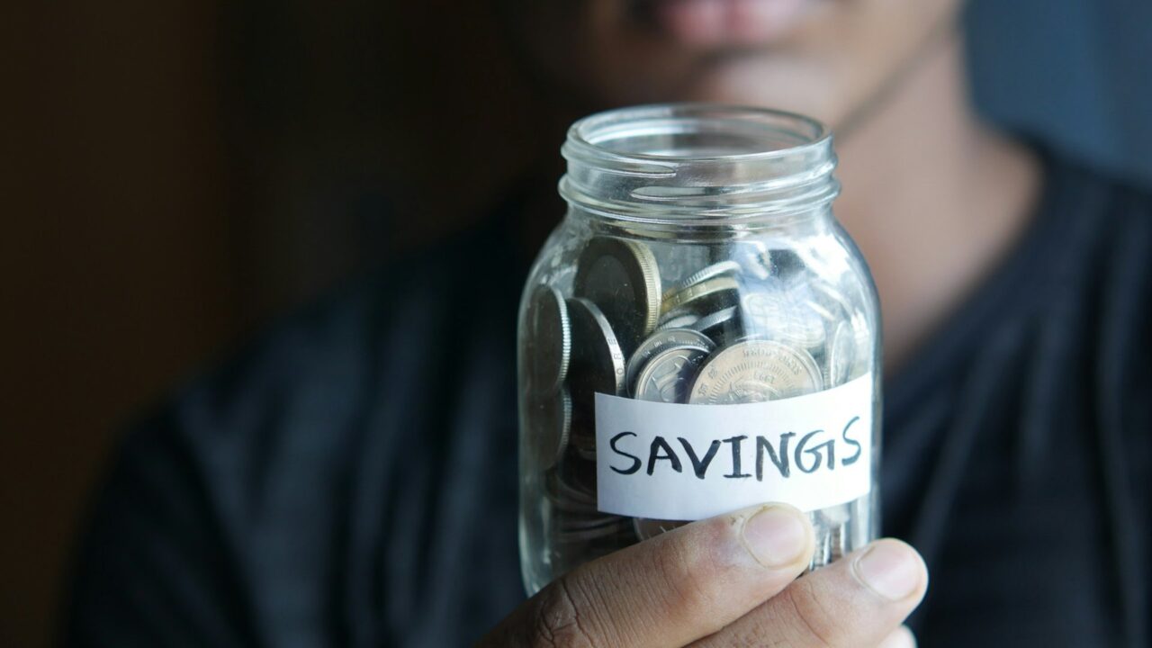 A person holding a clear glass jar filled with coins, with a 'Savings' label visible on it