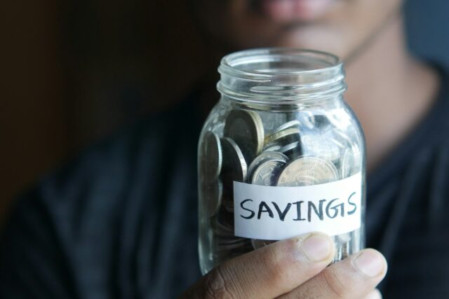 A person holding a clear glass jar filled with coins, with a 'Savings' label visible on it