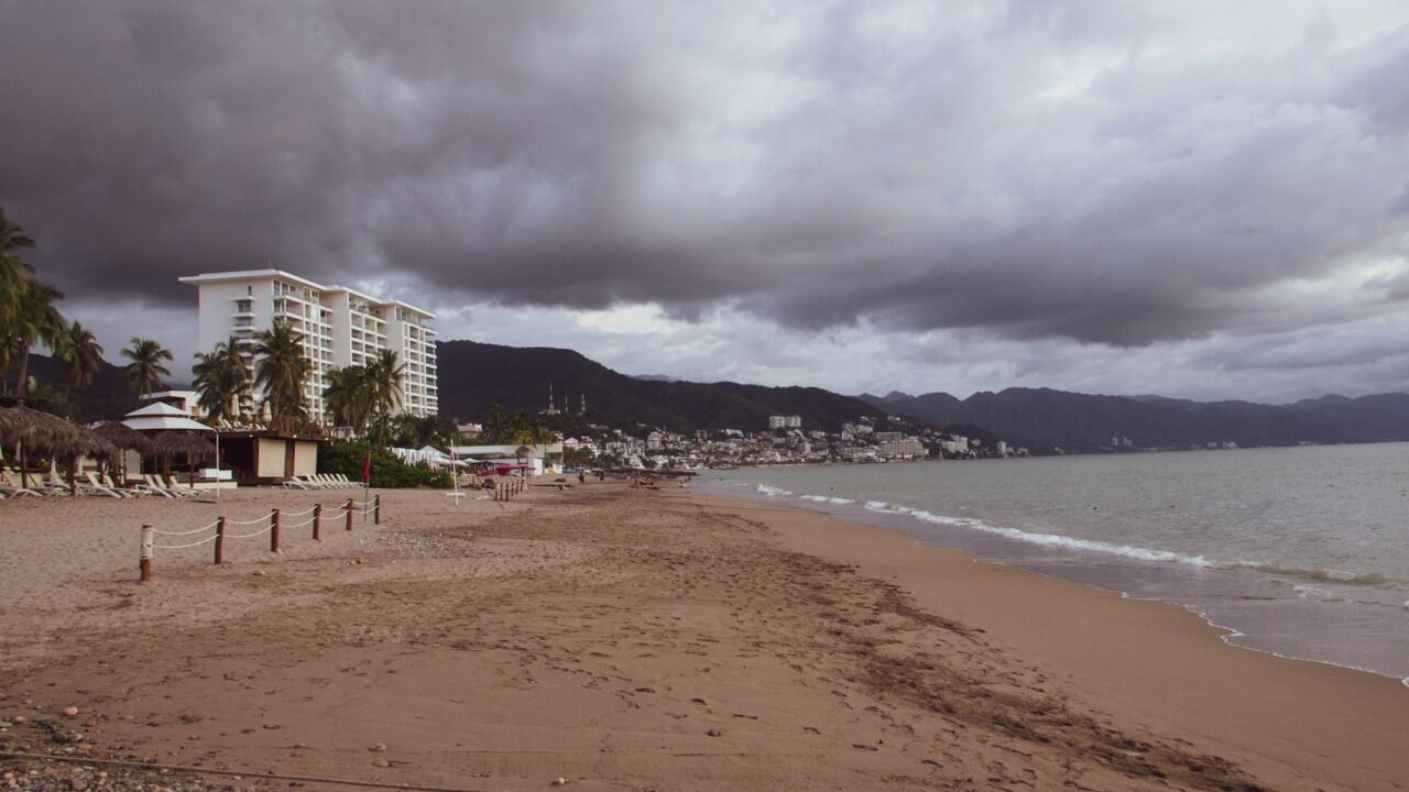A cloudy beach with dark storm clouds overhead, a hotel building, and mountains in the background during overcast weather conditions.