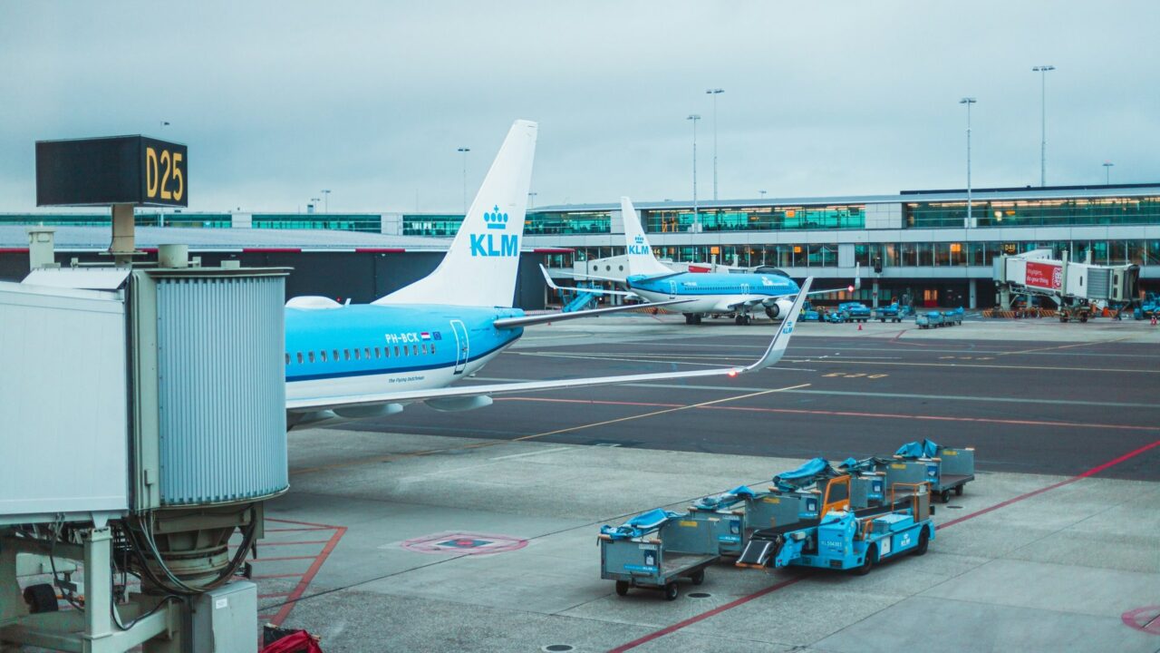A blue and white aircraft parked at a UK airport terminal with ground support vehicles visible