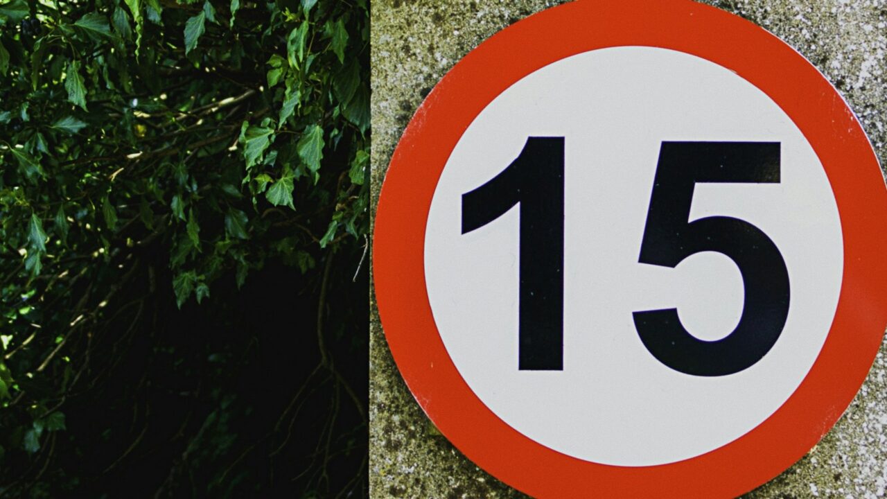 A red and white circular 15 km/h speed limit sign mounted on a concrete wall next to green foliage