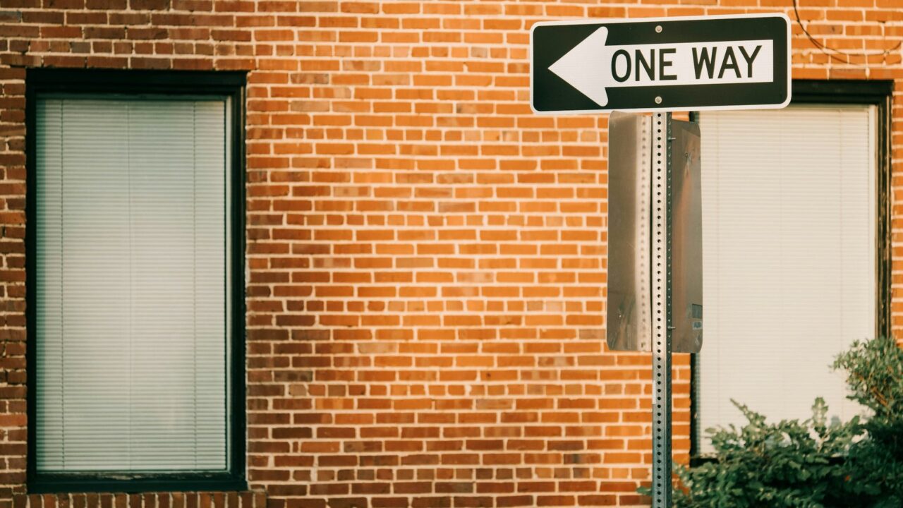 A black and white 'ONE WAY' street sign mounted on a red brick building with two doors