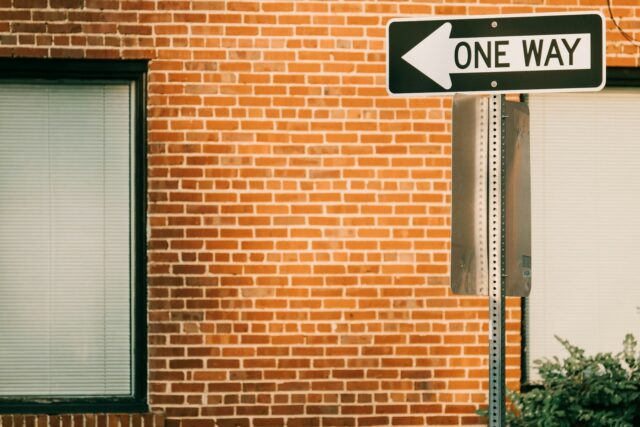 A black and white 'ONE WAY' street sign mounted on a red brick building with two doors