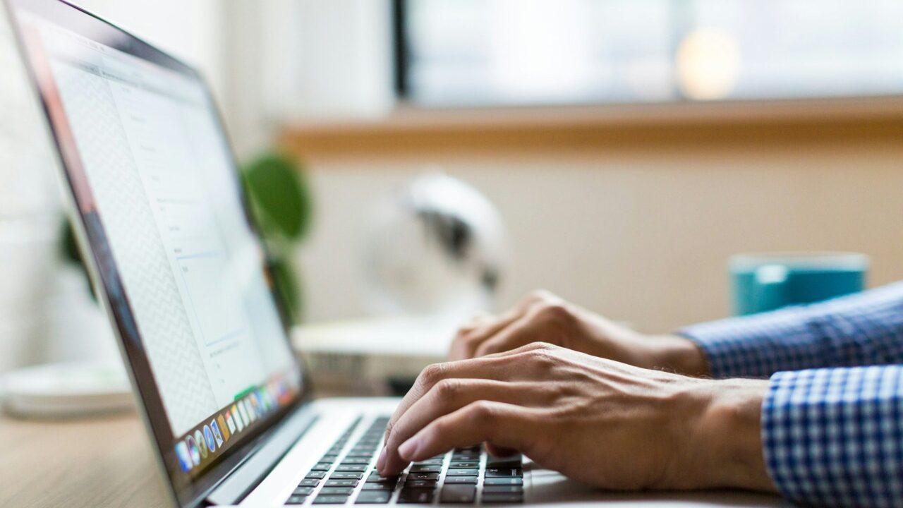 Person in checked shirt typing on a silver laptop at a desk in an office environment