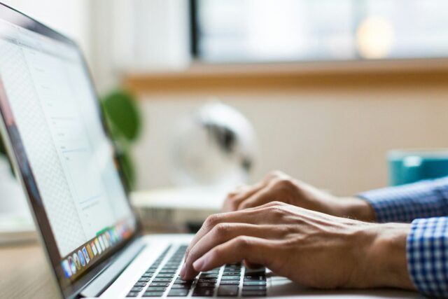 Person in checked shirt typing on a silver laptop at a desk in an office environment