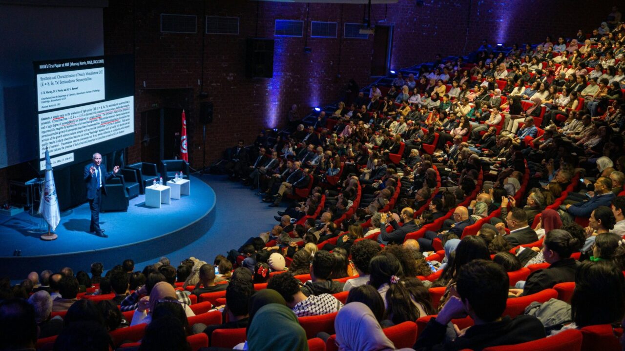 Speaker presenting to a large audience seated in an auditorium with purple stage lighting