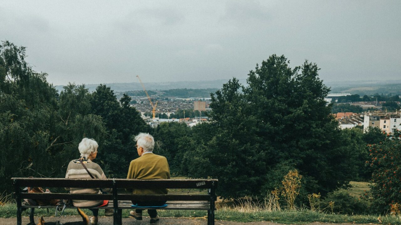 An elderly couple sitting on a bench overlooking a countryside landscape with a small dog beside them