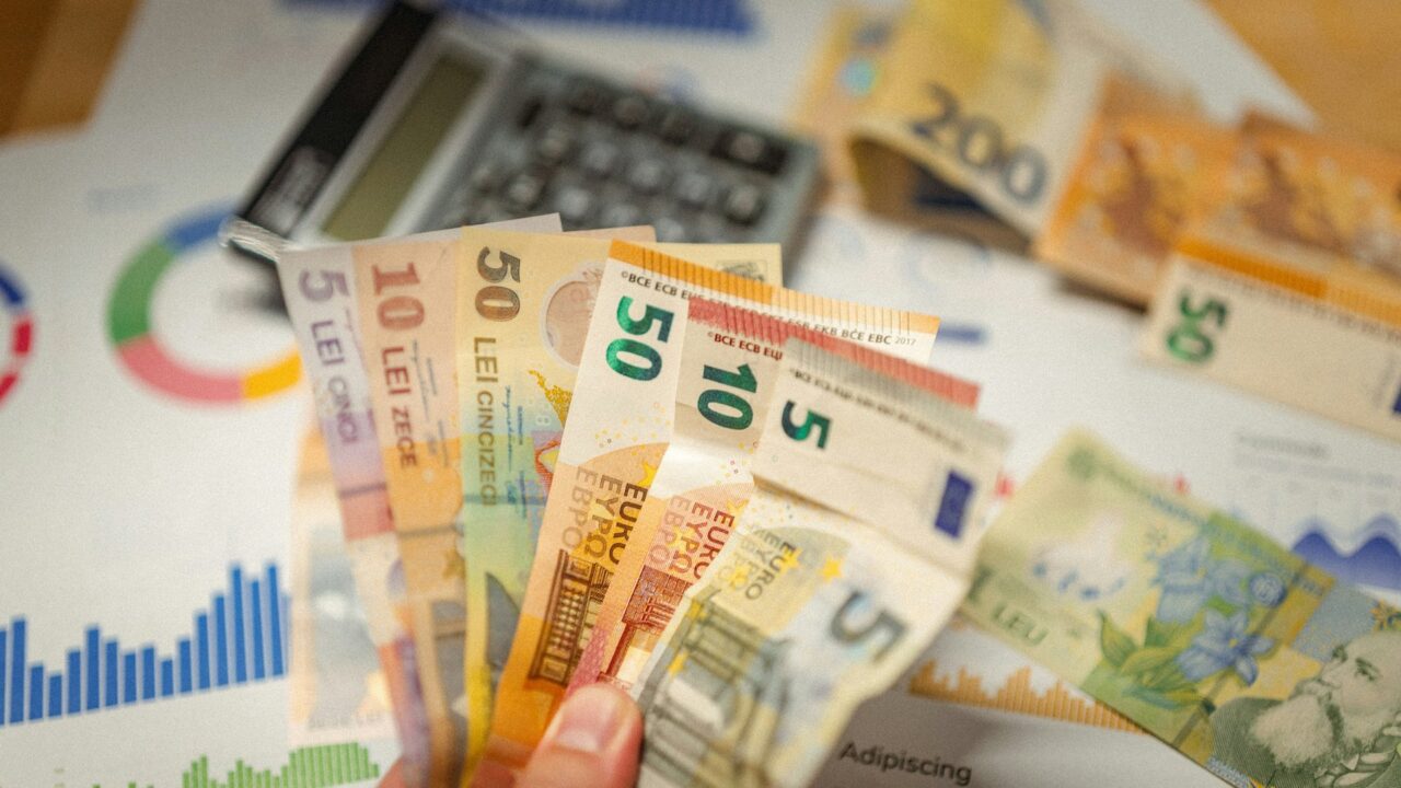 Hands holding Euro banknotes in front of a calculator and financial charts on a desk