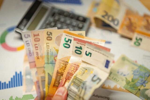 Hands holding Euro banknotes in front of a calculator and financial charts on a desk