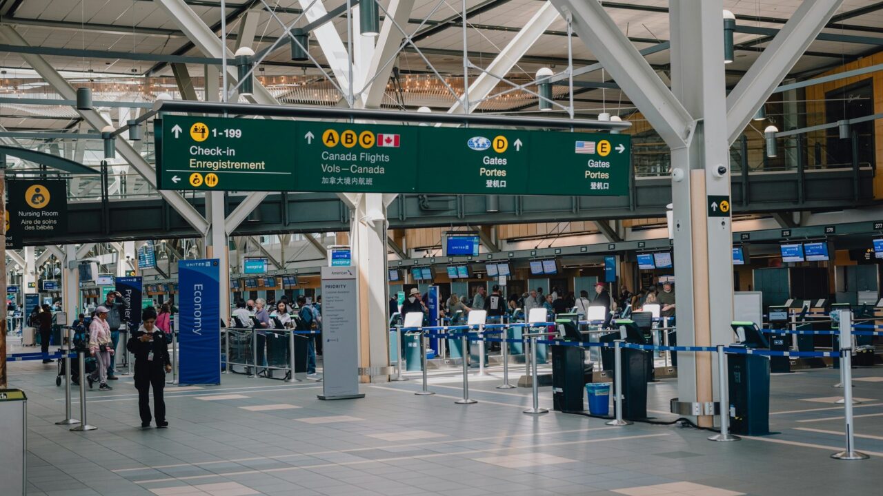 Interior of a UK airport terminal with overhead signage, departure boards, and passengers with luggage in the concourse area.