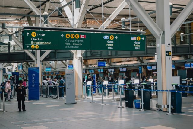 Interior of a UK airport terminal with overhead signage, departure boards, and passengers with luggage in the concourse area.