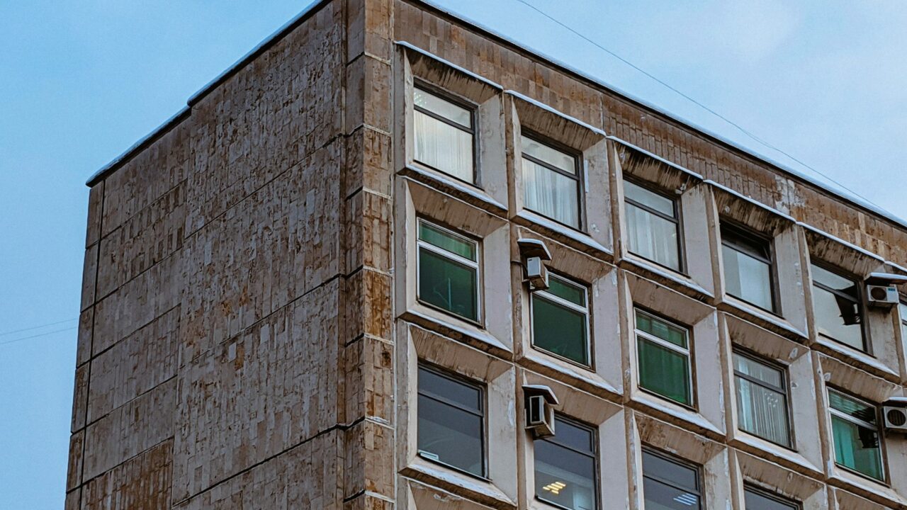 A multi-story residential building with weathered brown brick facade and regular window pattern showing signs of age and wear.