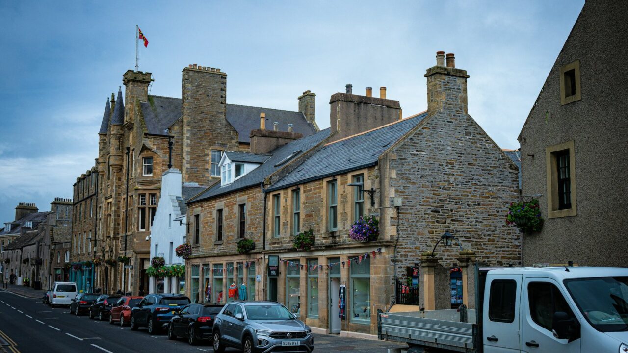 Stone buildings line a street with parked cars in a UK town
