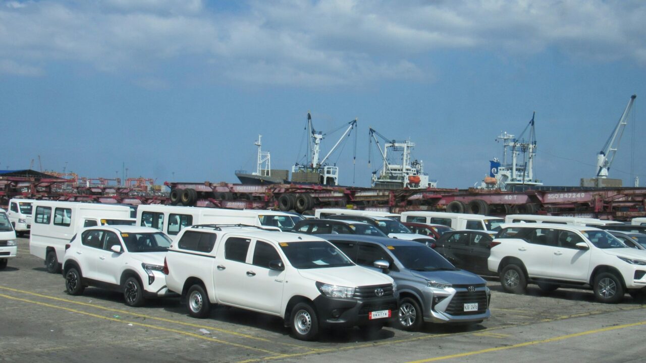 White vehicles parked at a port with cargo ships and cranes visible in the background