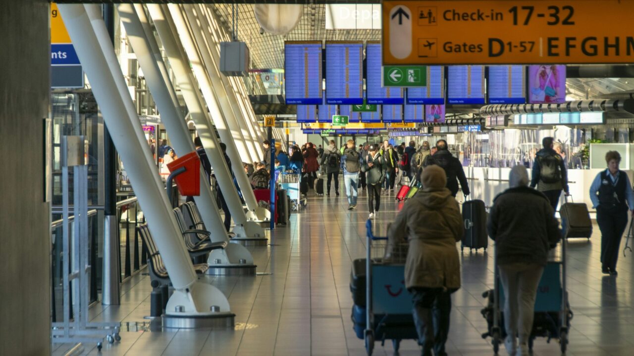 Travellers with luggage walking through an airport terminal with blue check-in gates and departure information signage visible overhead.