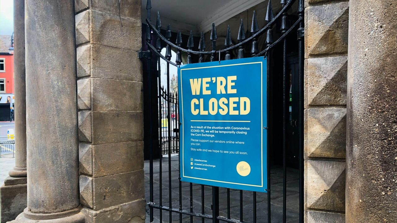 A blue and white 'We're Closed' sign displayed on black metal gates of a storefront.
