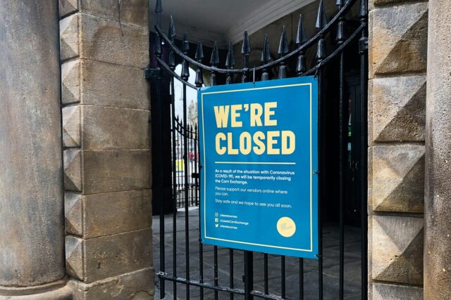 A blue and white 'We're Closed' sign displayed on black metal gates of a storefront.