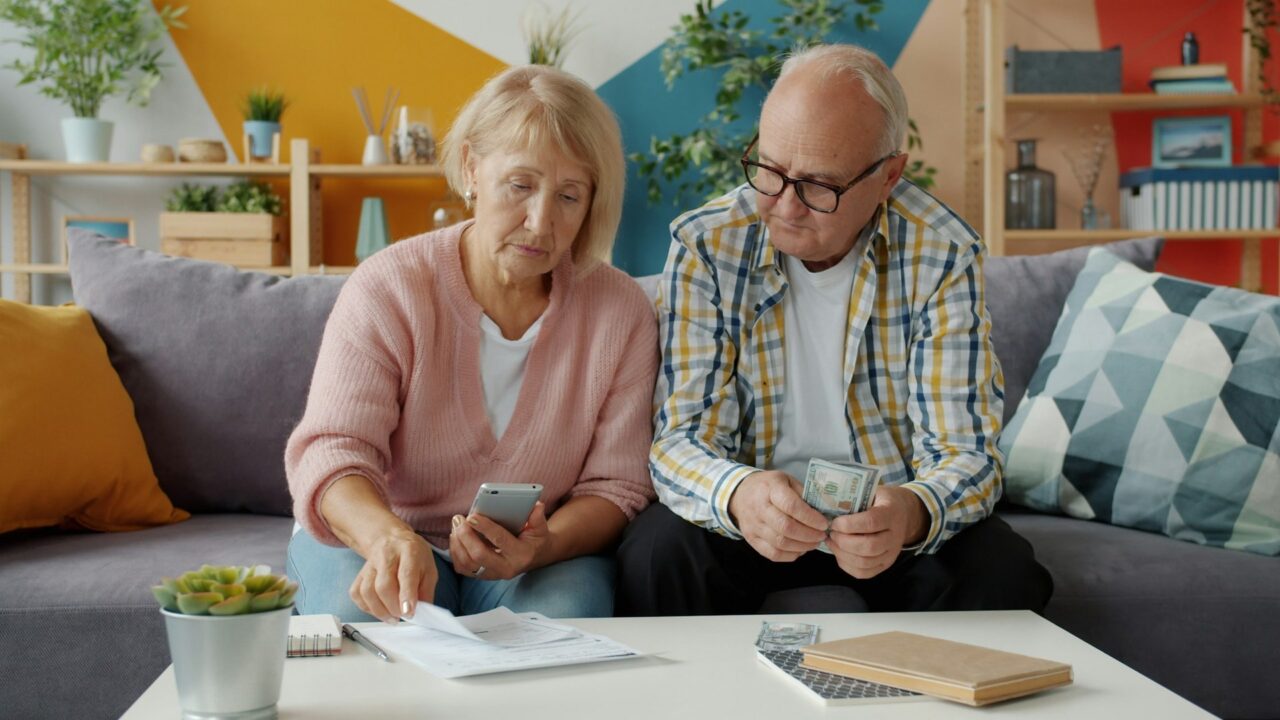 Elderly couple sitting on a sofa looking at bills and a mobile phone together in a home setting
